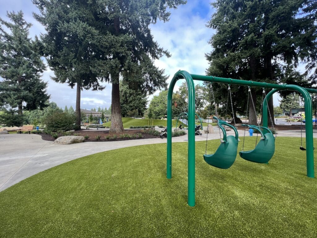 A green swingset with two swings facing a park with trees, walking paths, and play equipment.