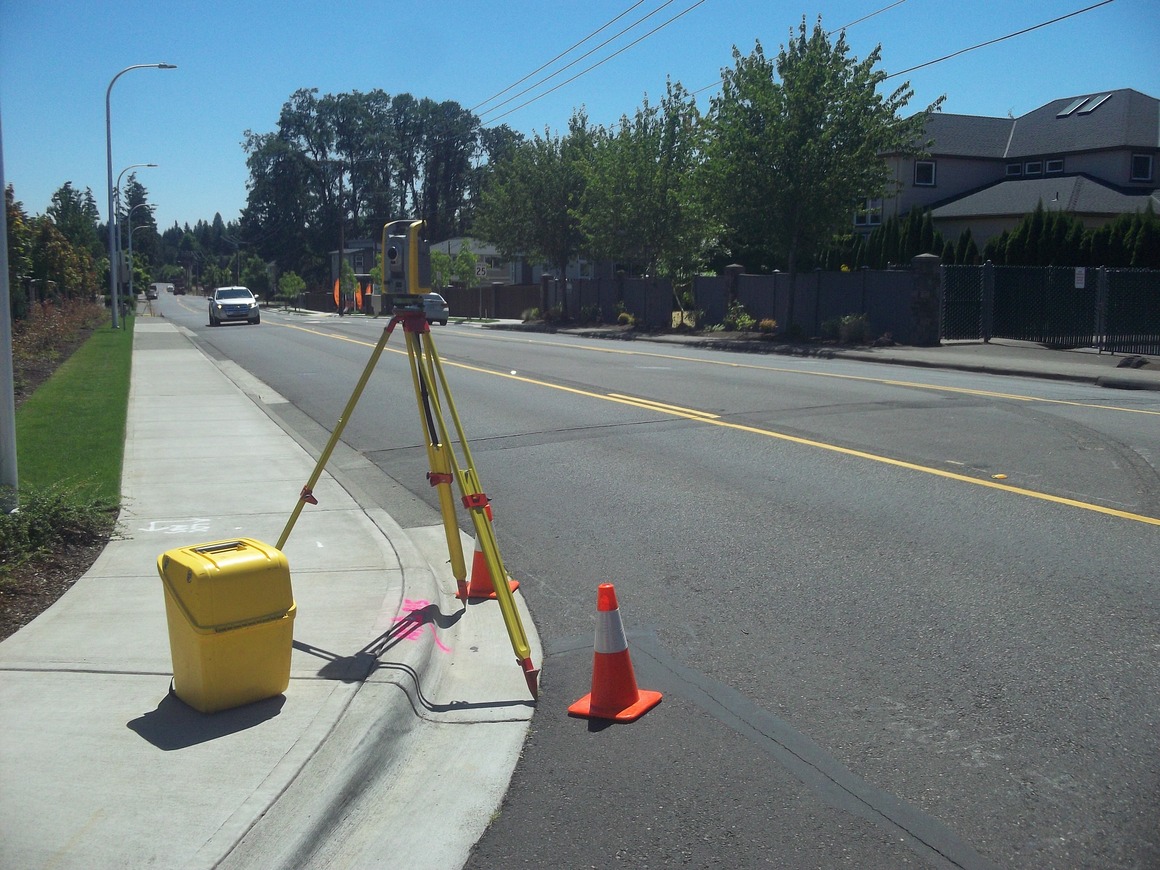 Photo of an orange traffic cone and total station set up on the sidewalk and street with a car in the distance