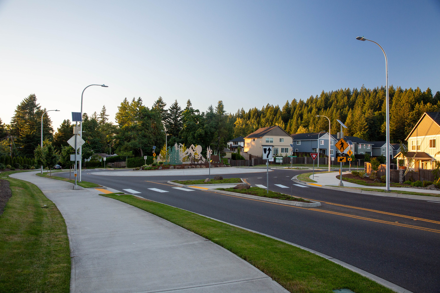 A neighborhood street with a crosswalk, pedestrian island at the intersection, a sidewalk and grass on the left, and houses to the upper right